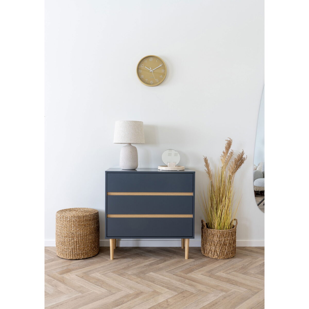In een moderne kamer staan een blauw dressoir met houten poten, een geweven krukje, een tafellamp, een decoratief object, een mand met gedroogd gras, een wandklok en een deel van de Table Mirror, allemaal op een vloer van visgraathout.