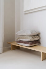 Een minimalistisch tafereel met het Mohair sierkussenhoes Amy lichtgrijs 50x50 cm gestapeld met andere neutrale kussens op een houten bank tegen een lichte muur in een lichte, moderne kamer.