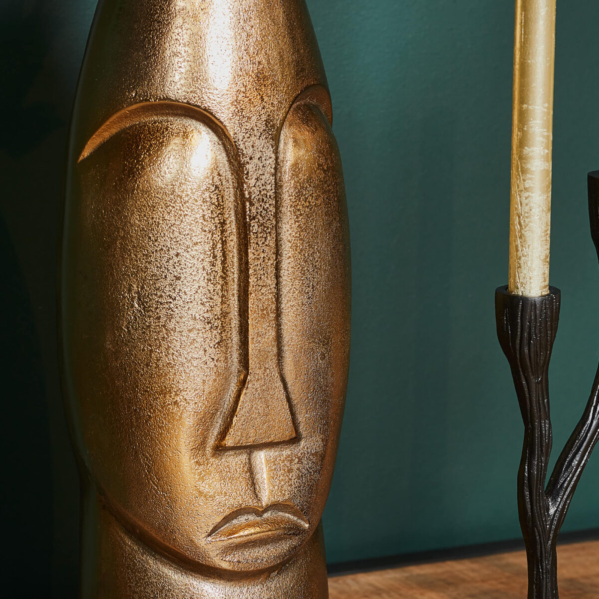 Een close-up van Vaas Lyra - Antiek goud | Large, een metallic gouden abstracte gezichtsvaas, naast een hoge zwarte kandelaar met een gele kaars, tegen een groene muur.