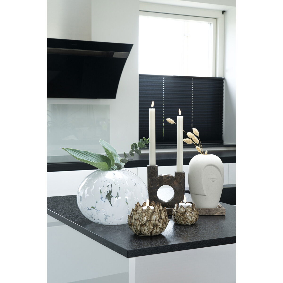 Op een modern aanrecht in natuurlijk licht staat de Ceramic Vase-vaas van zandkleurig keramiek met gezicht (12,5x13x19,5 cm)-met groene bladeren, plus gouden kaarsenhouders, een sculpturale kaarsenhouder met brandende kaarsen en een decoratief wit object in de vorm van een hoofd.
