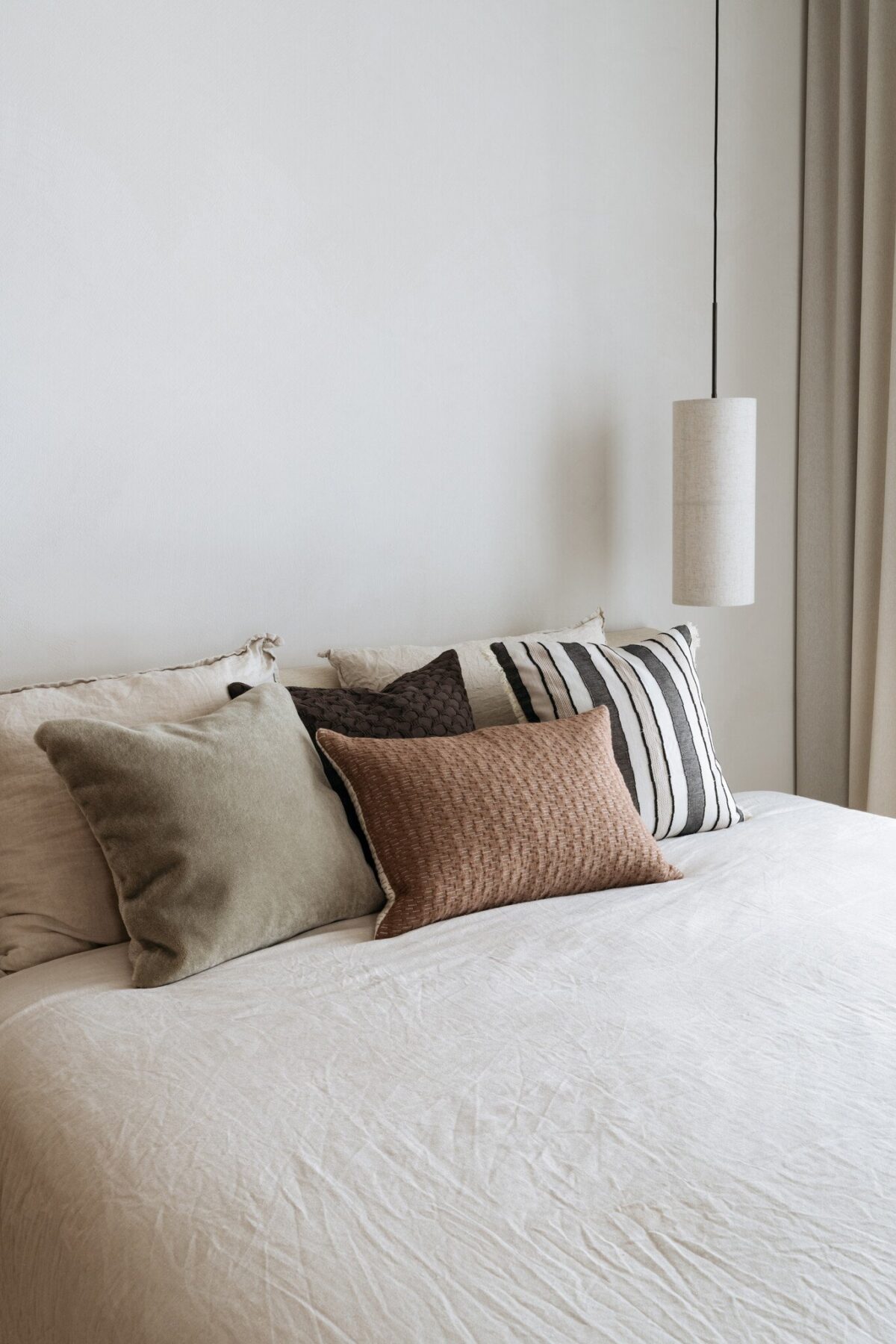 Op een keurig opgemaakt bed ligt het Mohair sierkussenhoes Amy grijsgroen 50x50 cm tussen neutrale kussens in beige en bruin, tegen een effen witte muur. Een cilindervormige hanglamp hangt erboven, met beige gordijnen gedeeltelijk zichtbaar aan de rechterkant.