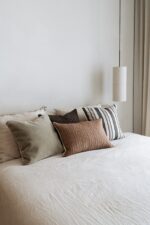 Op een keurig opgemaakt bed ligt het Mohair sierkussenhoes Amy grijsgroen 50x50 cm tussen neutrale kussens in beige en bruin, tegen een effen witte muur. Een cilindervormige hanglamp hangt erboven, met beige gordijnen gedeeltelijk zichtbaar aan de rechterkant.