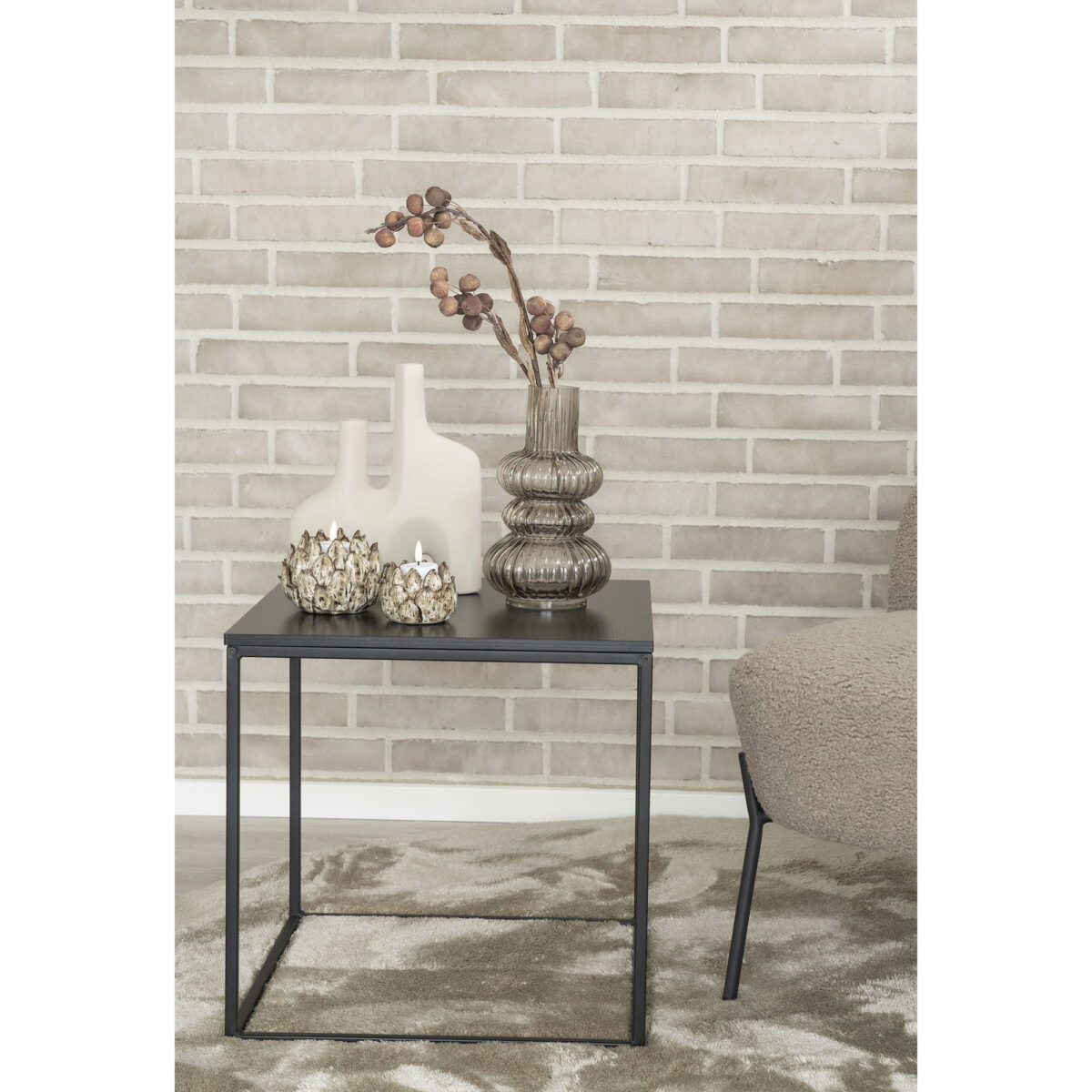 Een klein zwart metalen bijzettafeltje met een vaas van zandkleurig keramiek (twee openingen, 8x23,5x27 cm), gedroogde takken en twee kandelaars staat op een kleed naast een beige stoel tegen een lichte bakstenen muur.