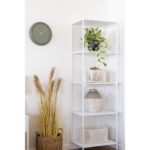Op een witte metalen plank staan een vaas, een bebladerde potplant en witte manden. Vlakbij staat een mand met hoog gedroogd gras. Boven de plank hangt een ronde groene wandklok aan de witte muur.