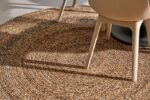 Een close-up van het Bellano ST Light Grey 814 ø200 rond geweven jute tapijt met twee lichtgekleurde moderne stoelen erop, hun schaduwen geworpen over het tapijt.
