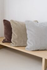 Het Mohair sierkussenhoes Amy lichtgrijs 50x50 cm staat op een eenvoudige houten bank, vergezeld van pluche kussens in bruin en beige, tegen een effen witte muur.