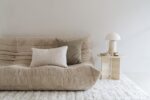 Een beige ribfluwelen sofa met twee Mohair sierkussenhoezen Amy grijsgroen 50x50 cm ligt op een wit getextureerd tapijt naast een kleine ronde tafel met een moderne witte lamp, een kopje en gebak tegen een effen witte muur.