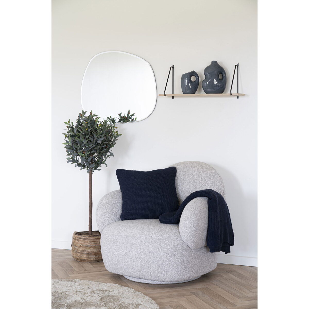 Een knusse fauteuil met een marineblauw kussen en een deken staat bij een plant in pot. Boven een houten plank staan twee vazen en aan de witte muur in deze lichte, minimalistische kamer hangt een onregelmatige spiegel.