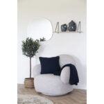 Een knusse fauteuil met een marineblauw kussen en een deken staat bij een plant in pot. Boven een houten plank staan twee vazen en aan de witte muur in deze lichte, minimalistische kamer hangt een onregelmatige spiegel.