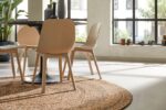 Moderne eethoek met Bellano ST Light Grey 814 ø200 ronde tafel met lichte houten stoelen op een geweven tapijt; grote ramen vullen de ruimte met natuurlijk licht en groene planten zorgen voor decoratieve accenten.