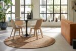 Moderne eethoek met een ronde tafel en drie stoelen op het tapijt Bellano ST Light Grey 814 ø200 bij een groot raam. Een bruine sofa en groene planten zijn zichtbaar, met binnenvallend zonlicht.