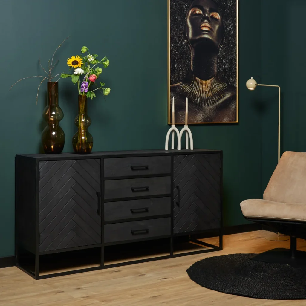 Een moderne kamer met donkergroene muren, het Dressoir New York | 165 cm zwarte dressoir met laden, twee hoge vazen met bloemen, een kandelaar, een vrouwenportret, een bruine loungestoel en een rond zwart vloerkleed op een houten vloer.
