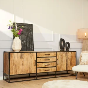 De Dressoir Denver (mangohout en staal, 210 cm) toont een witte vaas met kleurrijke bloemen, abstracte zwarte kunst en twee zwarte sculpturen, naast een beige stoel en een hoge staande lamp in een lichte, minimale kamer.