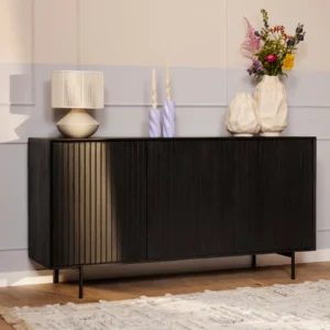 De Dressoir Madison | 165 cm, een modern zwart dressoir met verticale groeven, staat tegen een lichte muur met daarop een geribbelde lamp, twee golvende kaarsen, twee witte vazen en een kleurrijk boeket. Een wit vloerkleed is gedeeltelijk zichtbaar op de vloer.