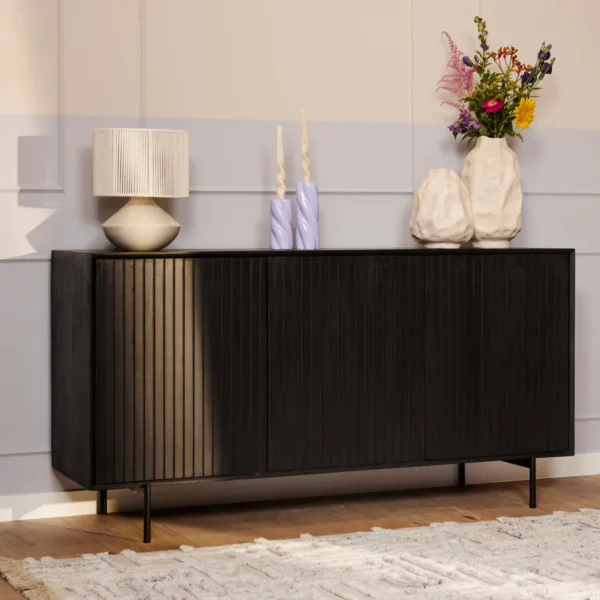 De Dressoir Madison | 165 cm, een modern zwart dressoir met verticale groeven, staat tegen een lichte muur met daarop een geribbelde lamp, twee golvende kaarsen, twee witte vazen en een kleurrijk boeket. Een wit vloerkleed is gedeeltelijk zichtbaar op de vloer.