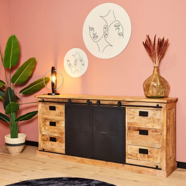 De Dressoir Fresno 200 cm met zwarte schuifdeuren en laden staat tegen een roze muur, met daarop een potplant, lamp en vaas met droogbloemen. Erboven hangen twee minimalistische lijnkunstportretten.