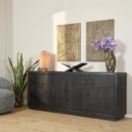 De Dressoir Brussel Zwart 4 deurs 200 cm is ingericht met een roze lamp, sculpturaal decor en een gouden vaas met paarse bloemen. Vlakbij staat een hoge groene plant, met daarboven twee abstracte gouden kunstwerken aan de muur.