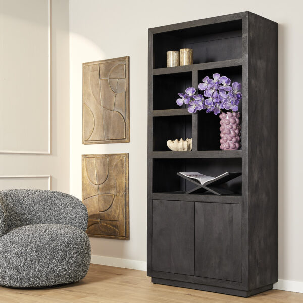 De Boekenkast Brussel Zwart met 5 planken en 2 deuren staat tegen een witte muur met boeken, decor en een vaas paarse bloemen naast een fauteuil met grijze structuur en abstracte muurkunst.