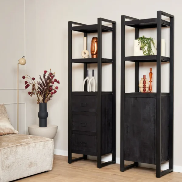 Twee Boekenkast Boaz Black boekenkasten met deuren, versierd met vazen, planten en beelden, staan tegen een beige muur bij een crèmekleurige bank en een staande lamp met een vaas met rode en paarse bladeren.