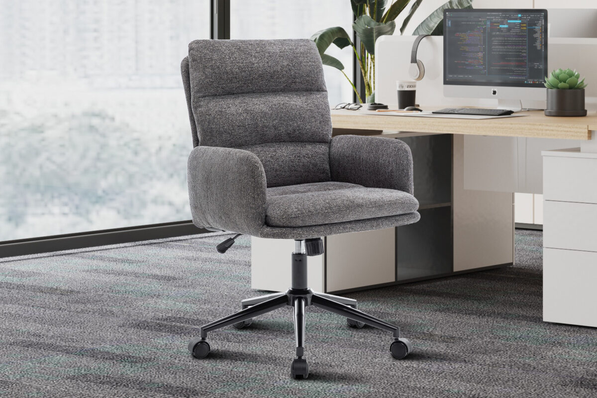 De Bureaustoel 'Aimé' Stof Fabb Raven 13 Donkergrijs ligt op een vloerbedekking naast een houten bureau met een computer, toetsenbord, plant en decor in een lichte kantoorruimte.