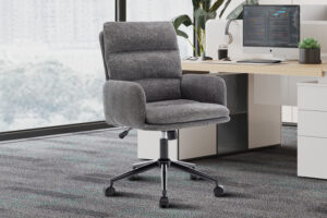 De Bureaustoel 'Aimé' Stof Fabb Raven 13 Donkergrijs ligt op een vloerbedekking naast een houten bureau met een computer, toetsenbord, plant en decor in een lichte kantoorruimte.