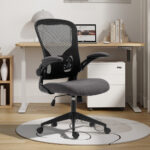 De Bureaustoel 'Marcos' Mesh Zwart/Grijs (1c), met een rugleuning van netstof en een gevoerde zitting op wieltjes, staat op een ronde mat bij een houten bureau met een laptop, lamp, boeken en decor in een licht, modern thuiskantoor.