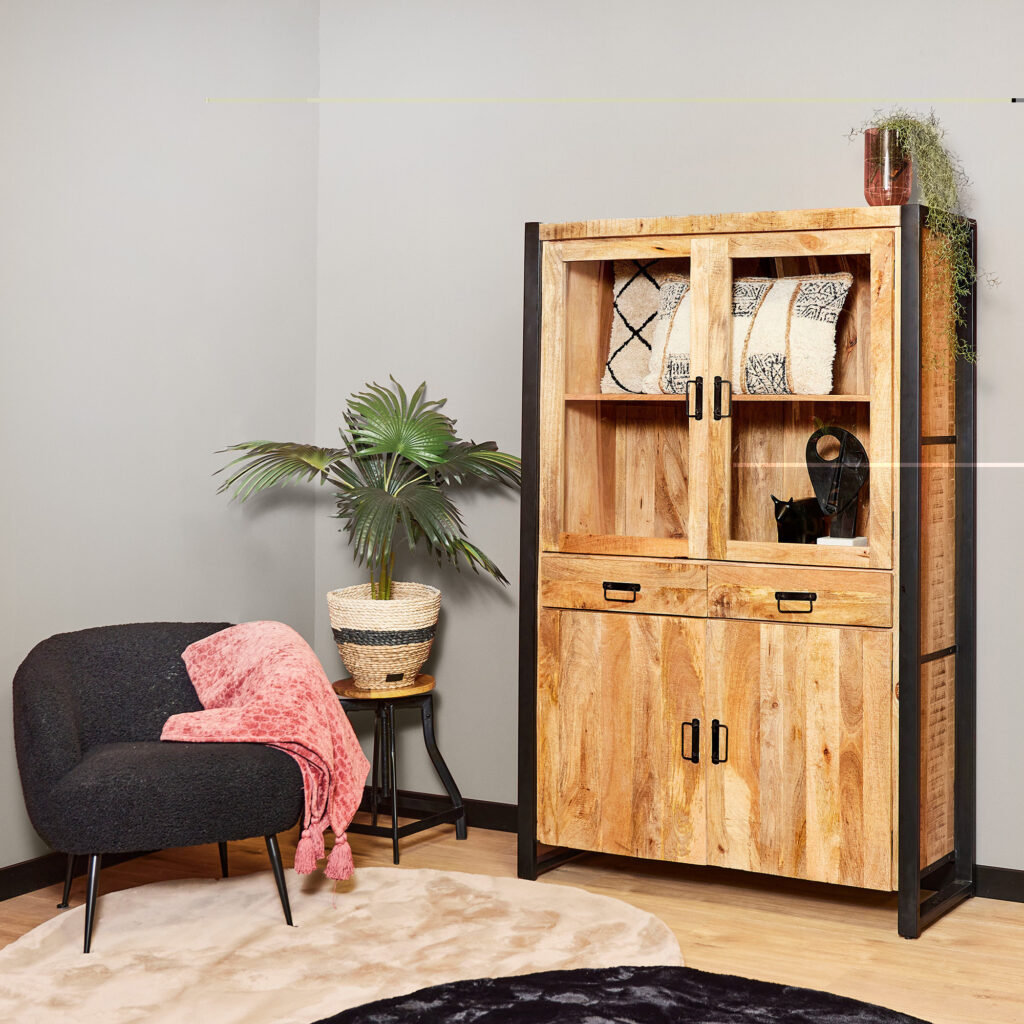 Een gezellige kamerhoek met een zwarte fauteuil en roze deken, een ronde zwarte bijzettafel met een palm in pot en de Vitrinekast Boaz 115 cm kast met daarop gedessineerde dekens, een vaas en een plant.