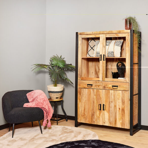 Een gezellige kamerhoek met een zwarte fauteuil en roze deken, een ronde zwarte bijzettafel met een palm in pot en de Vitrinekast Boaz 115 cm kast met daarop gedessineerde dekens, een vaas en een plant.