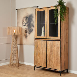 De Vitrinekast RichWood | 100 cm met glazen deuren staat naast een hoge rieten vloerlamp en wandkunstwerk van een gedeeltelijk verborgen gezicht. In de kast zit een klein wit boomdecor; bovenop zit een groene plant.