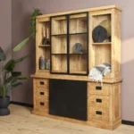 De Nashville Vitrinekast | 200 cm is een grote houten kast met lades, zwarte schuifdeuren en glazen bovenplanken om decor in uit te stallen. De kast wordt getoond naast een groene potplant in een moderne kamer met mauve muren en houten vloeren.