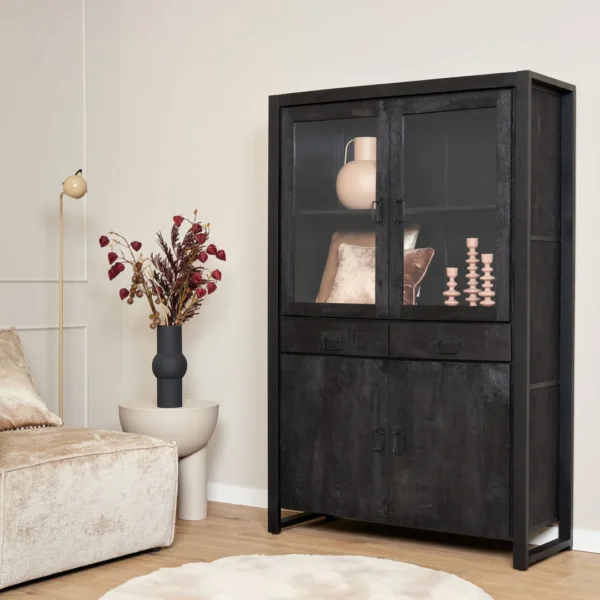 Een moderne woonkamerhoek met een beige bank, een ronde bijzettafel met droogbloemen, een hoge staande lamp en de Vitrinekast Boaz Black | 115 cm vitrinekast met glazen deuren waarin roze decoratieve items worden tentoongesteld.