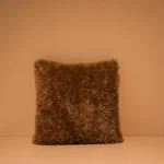 Het Armkussen 50x50, een vierkant kussen met bruine shaggy, staat rechtop tegen een beige muur op een wit vlak, verlicht met warm licht.