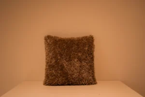 Het Armkussen 50x50, een vierkant kussen met bruine shaggy, staat rechtop tegen een beige muur op een wit vlak, verlicht met warm licht.