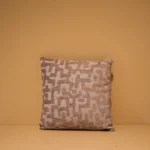 Het Armkussen 50x50, een vierkant sierkussen in beige en crème met een gestructureerd geometrisch patroon, is gecentreerd tegen een effen mosterdgele achtergrond.