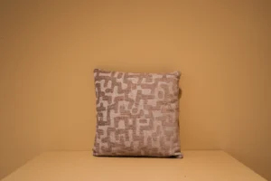 Het Armkussen 50x50, een vierkant sierkussen in beige en crème met een gestructureerd geometrisch patroon, is gecentreerd tegen een effen mosterdgele achtergrond.