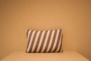 Het Armkussen 50x50, een rechthoekig kussen met bruine en witte diagonale strepen, rust op een beige ondergrond met een effen beige muur op de achtergrond.