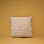 Het Armkussen 50x50, een vierkant kussen met een geometrisch patroon in beige en lichtgrijs, is gecentreerd tegen een effen bruine achtergrond en oppervlak.