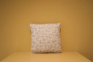 Het Armkussen 50x50, een vierkant kussen met een geometrisch patroon in beige en lichtgrijs, is gecentreerd tegen een effen bruine achtergrond en oppervlak.