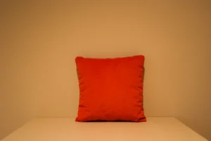 Het Armkussen 50x50, een levendig oranje vierkant kussen, is gecentreerd op een effen wit oppervlak met een beige muur op de achtergrond voor een strakke, minimalistische look.