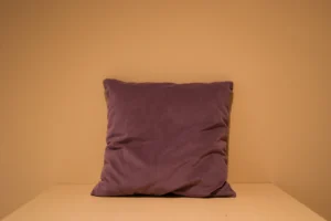 Het Armkussen 50x50, een paars fluwelen kussen, staat rechtop tegen een effen beige muur op een bijpassend oppervlak. Het zacht verlichte en minimalistische tafereel benadrukt het elegante ontwerp.