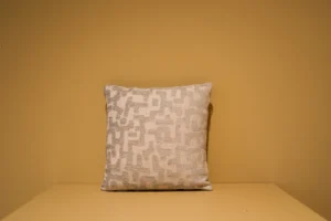 Het Armkussen 50x50, een vierkant kussen met een lichtbeige en wit geometrisch patroon, ligt tegen een mosterdgele muur op een bijpassend geel vlak.