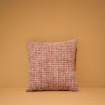 Het Armkussen 50x50, een vierkant kussen met een oranje en wit geweven structuurpatroon, ligt op een vlakke ondergrond voor een effen lichtbruine muur.
