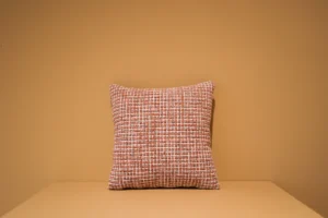 Het Armkussen 50x50, een vierkant kussen met een oranje en wit geweven structuurpatroon, ligt op een vlakke ondergrond voor een effen lichtbruine muur.
