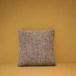 Het Armkussen 50x50, een vierkant kussen met een zwart-wit geblokt patroon, is tentoongesteld op een vlak beige oppervlak tegen een bijpassende beige muur.