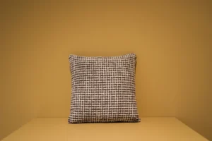 Het Armkussen 50x50, een vierkant kussen met een zwart-wit geblokt patroon, is tentoongesteld op een vlak beige oppervlak tegen een bijpassende beige muur.