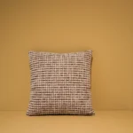 Het Armkussen 50x50, een vierkant kussen met een geweven ruitpatroon in bruin, wit en zwart, rust tegen een effen lichtbruine muur op een bijpassend oppervlak.