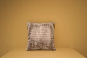 Het Armkussen 50x50, een vierkant kussen met een geweven ruitpatroon in bruin, wit en zwart, rust tegen een effen lichtbruine muur op een bijpassend oppervlak.