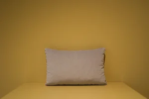 Het Armkussen 50x50, een effen lichtbeige rechthoekig kussen, rust tegen een effen mosterdgele muur en ondergrond, waardoor een eenvoudig en minimalistisch tafereel ontstaat.