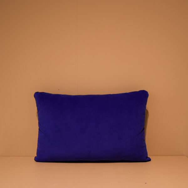 Het Armkussen 50x50, een rechthoekig diepblauw kussen, staat rechtop tegen een effen beige muur op een lichtbeige ondergrond in een eenvoudige en opgeruimde omgeving.