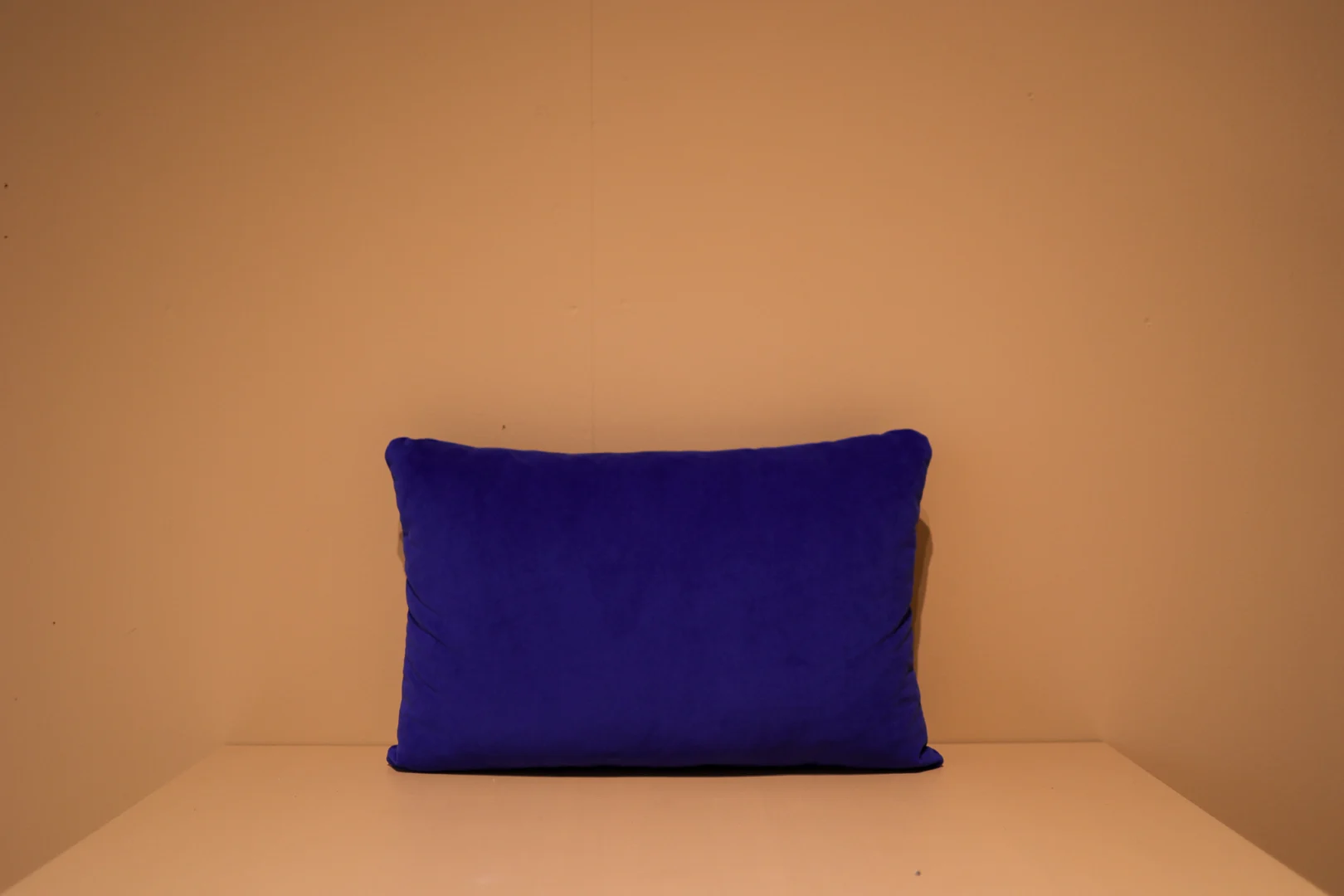 Het Armkussen 50x50, een rechthoekig diepblauw kussen, staat rechtop tegen een effen beige muur op een lichtbeige ondergrond in een eenvoudige en opgeruimde omgeving.